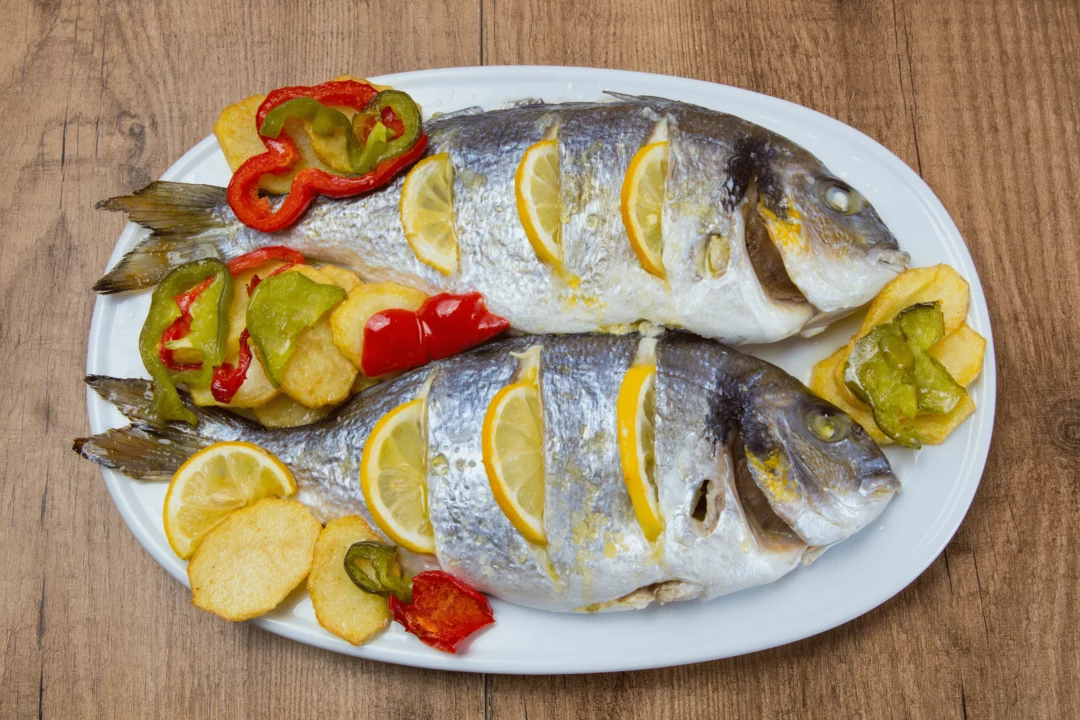 pecena ryba dorada s platky citronu a opecenymi bramborami podavana s paprikou na bilem taliri