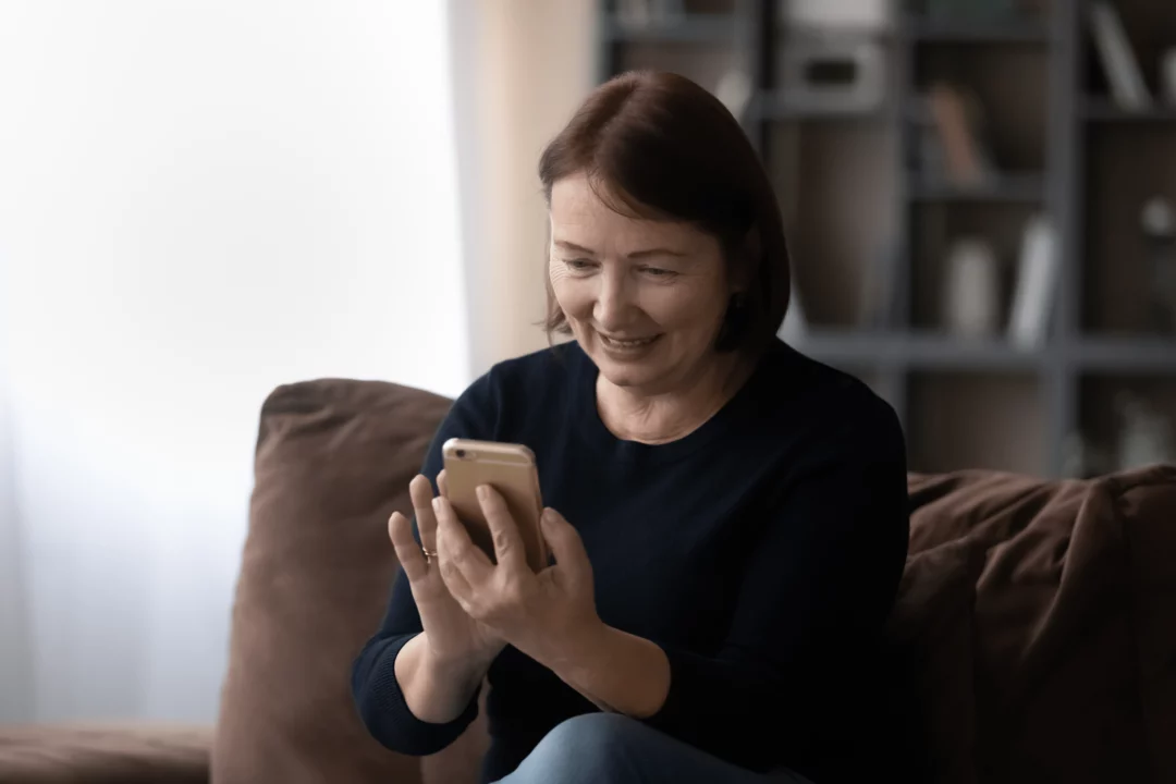 starsi zena sedici na gauci a usmivajici se zatimco pouziva chytry telefon v pohodli domova