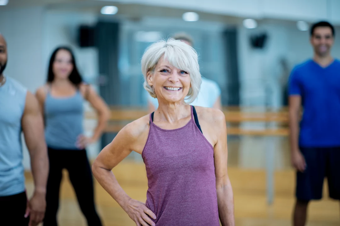 usmevava zena ve sportovnim obleceni pri skupinovem fitness cviceni ve svetlem studiu s nekolika lidmi v pozadi