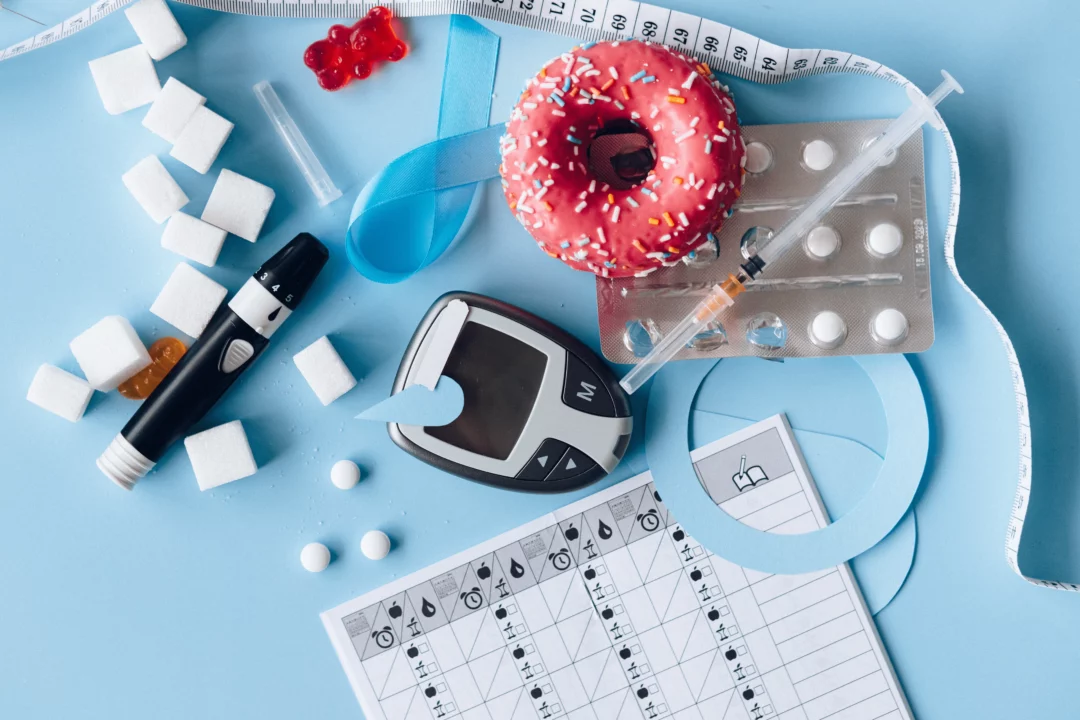 Diabetes koncept: glukometr, cukrové kostky, donuty, inzulínová stříkačka a dietní plán na modrém pozadí.