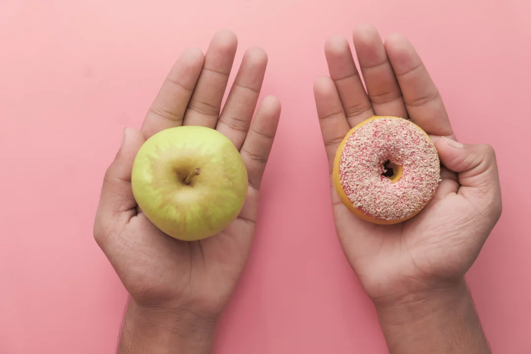 ruka drzici zelene jablko a druha ruka donut s polevou na ruzovem pozadi symbolizujici zdravou a nezdravou volbu