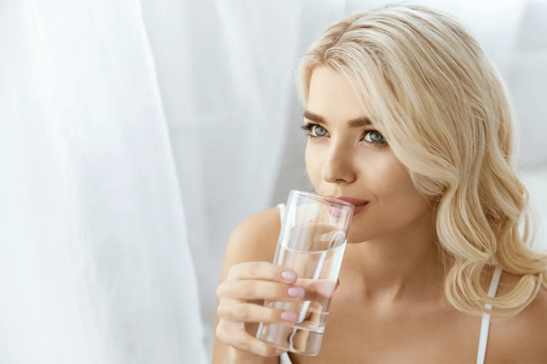 Žena s blond vlasy pije sklenici čisté vody, zdravý životní styl, hydratace, světlé pozadí.