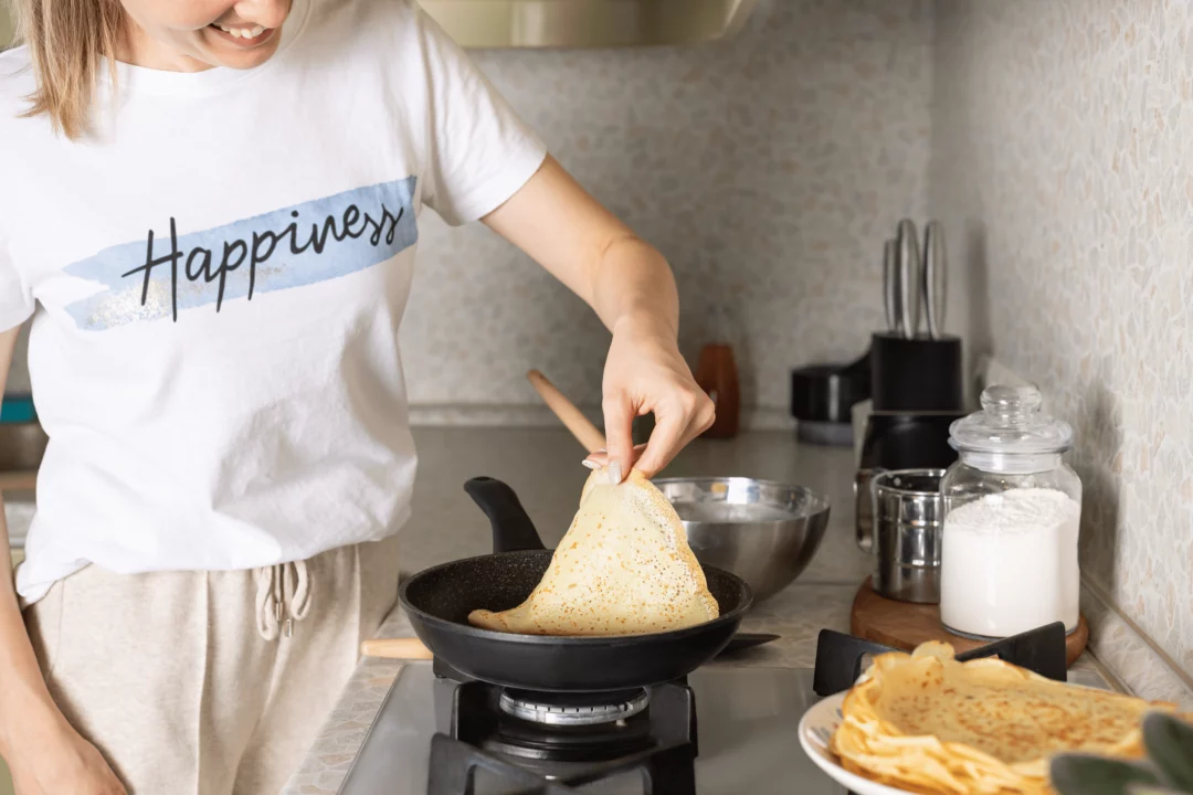 osoba smazi palacinky na panvi v kuchyni oblecena v tricku s napisem happiness na stole mouka a talir