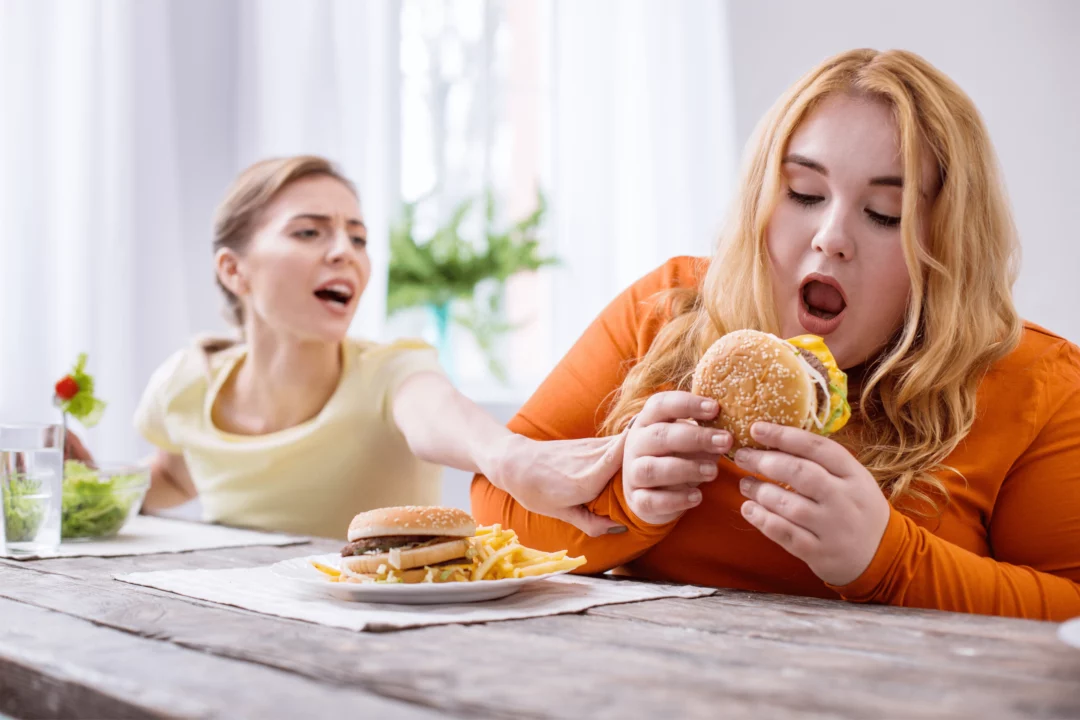 zena v oranzovem drzi burger zatimco druha zena se snazi zabranit v konzumaci nezdraveho jidla