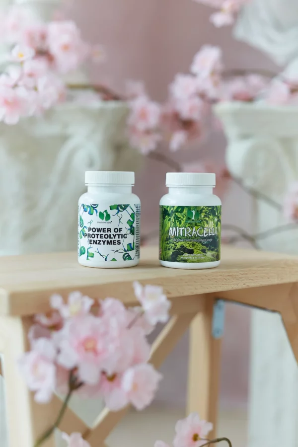 Dvě lahvičky doplňků stravy s označením „Power of Proteolytic Enzymes“ a „Mitracell“ na dřevěném povrchu s růžovými květy a rozmazaným pozadím.