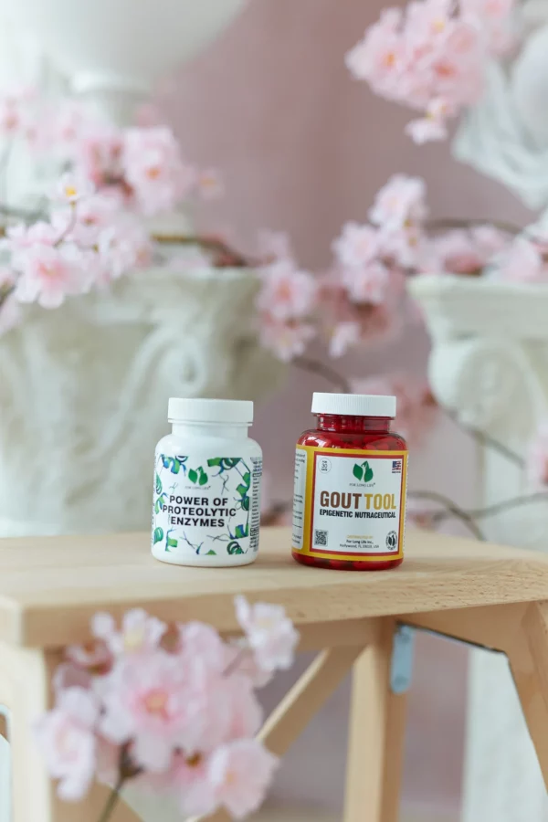 Dvě lahvičky doplňků stravy, „Power of Proteolytic Enzymes“ a „Gout Tool“, vystavené na dřevěném stole obklopeném růžovými třešňovými květy.