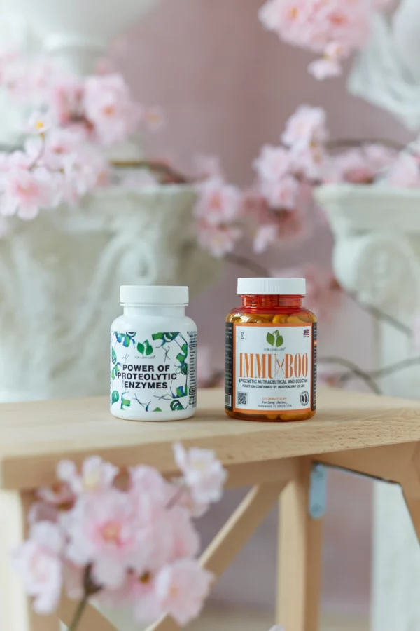 Dvě lahvičky doplňků stravy, „Power of Proteolytic Enzymes“ a „IMMUBOO“, na dřevěném povrchu obklopeném růžovými květy třešňového květu.