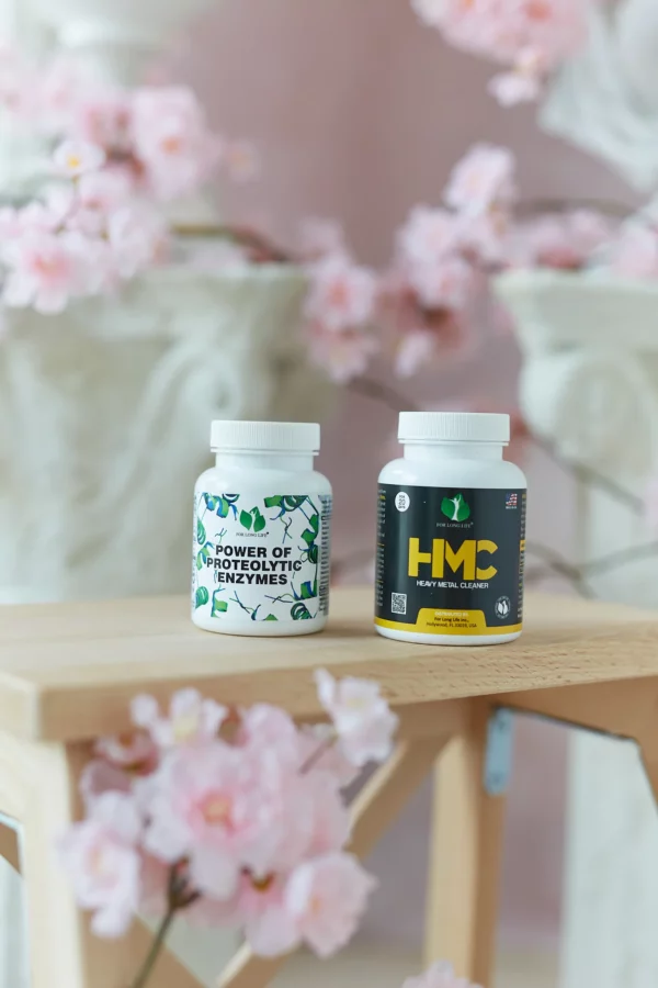 Na stole jsou vystaveny dvě lahvičky Power of Proteolytic Enzymes a HMV vedle zářivě aranžované růžové květiny.